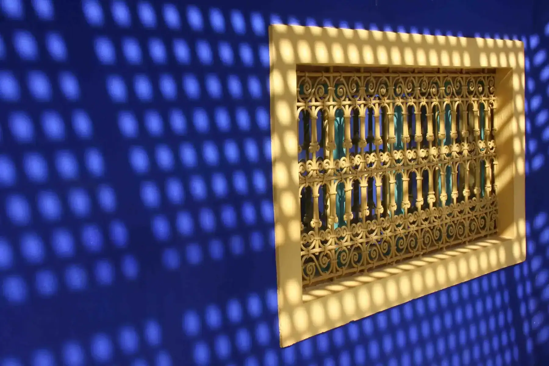 ljvdbos0-window-1207159_1920 Blue wall with a yellow framed ornate iron grille window; dotted shadow pattern covers the wall created by the grille's shapes.