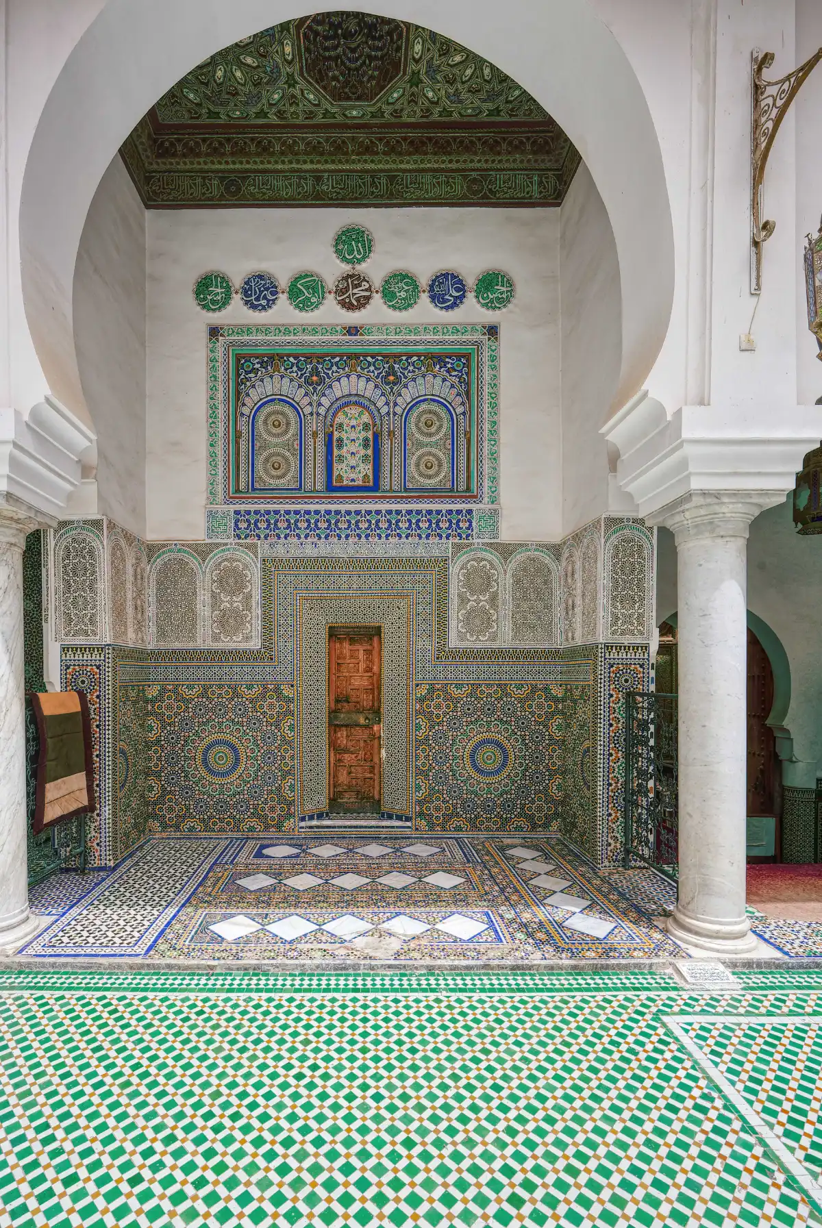 interieur-mosquee-moulay-idriss-fes-zellige Patio intérieur de la mosquée Moulay Idriss à Fès avec murs en zelliges multicolores, colonnes en marbre blanc et calligraphie arabe ornementale.