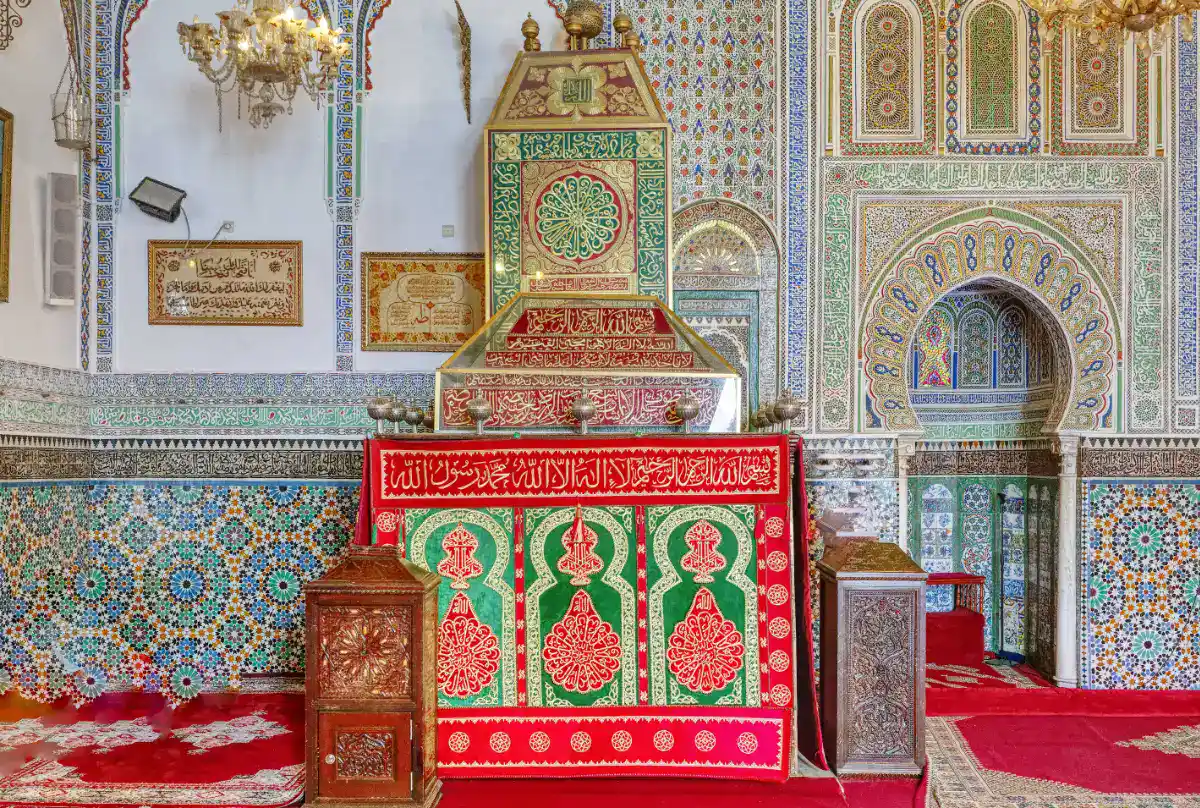Intérieur du mausolée de Moulay Idriss II à Fès montrant les murs en zelliges colorés, la calligraphie arabe et le catafalque recouvert d'un tissu vert et rouge.