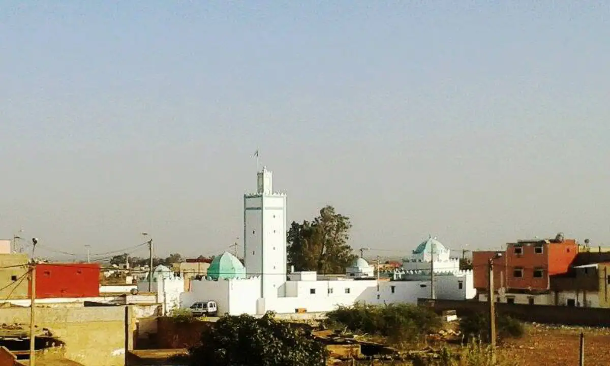 Desert town skyline with white buildings and a tall white minaret next to a green-domed mosque under a clear sky