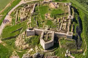Photographie en plongée des vestiges archéologiques en pierre du château d'Alarcos situés sur une colline verdoyante. Le site dévoile un vaste réseau de fondations anciennes, des pans de murs partiels et, au premier plan, une imposante base de tour défensive de forme polygonale. Quelques minuscules silhouettes humaines sur le chemin de terre en haut à gauche permettent d'apprécier la vaste échelle de ces ruines médiévales.