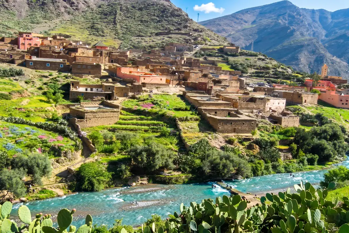 Paysage de montagne du Haut Atlas montrant un village traditionnel aux maisons en terre et en pierre ocre, bâti à flanc de colline verdoyante au-dessus d'une rivière aux eaux turquoise bordée de cactus et de végétation.