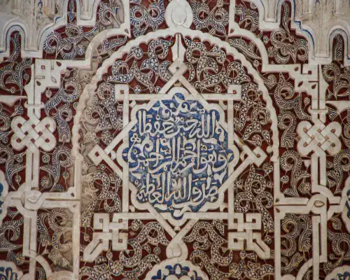 Art de la Calligraphie Andalouse Détail d'une calligraphie arabe sculptée sur stuc avec des motifs d'entrelacs et de l'écriture bleue au centre, typique de l'architecture nasride en Espagne.