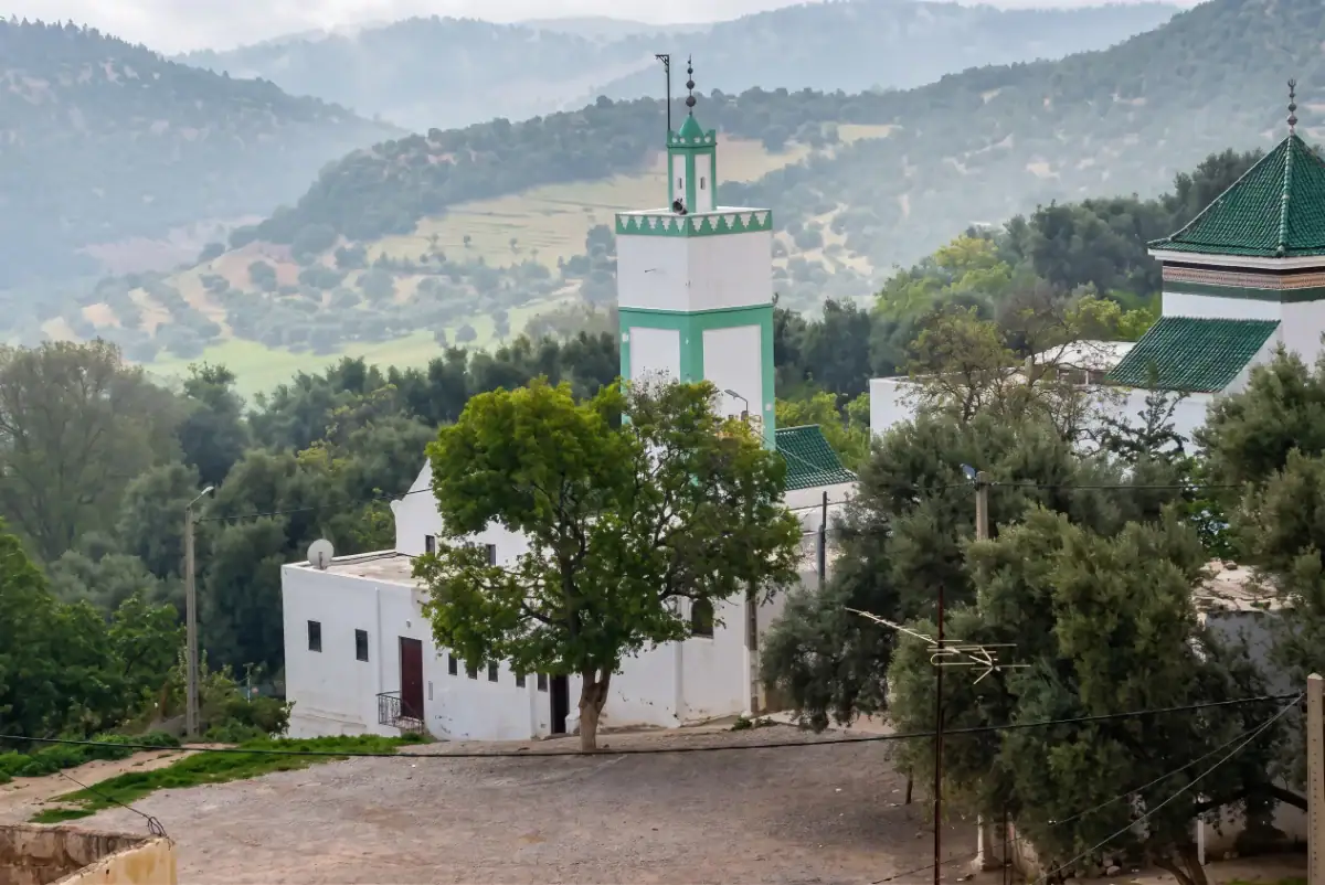 Mausolée de Sidi Lahcen Lyoussi Vue sur le mausolée de Sidi Lahcen Lyoussi avec son minaret blanc et vert, niché au cœur d'un paysage de collines verdoyantes.