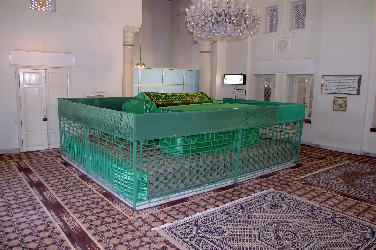 Vue de l'intérieur du mausolée de Cheikh Ahmed Zarrouq Al Fassi, montrant le tombeau recouvert d'un drap vert brodé, entouré d'une grille de protection verte et de tapis traditionnels.