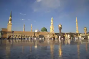 Vue de la Mosquée du Prophète Al-Masjid an-Nabawi à Médine avec son Dôme Vert emblématique et ses minarets, reflétée sur le sol de marbre blanc.