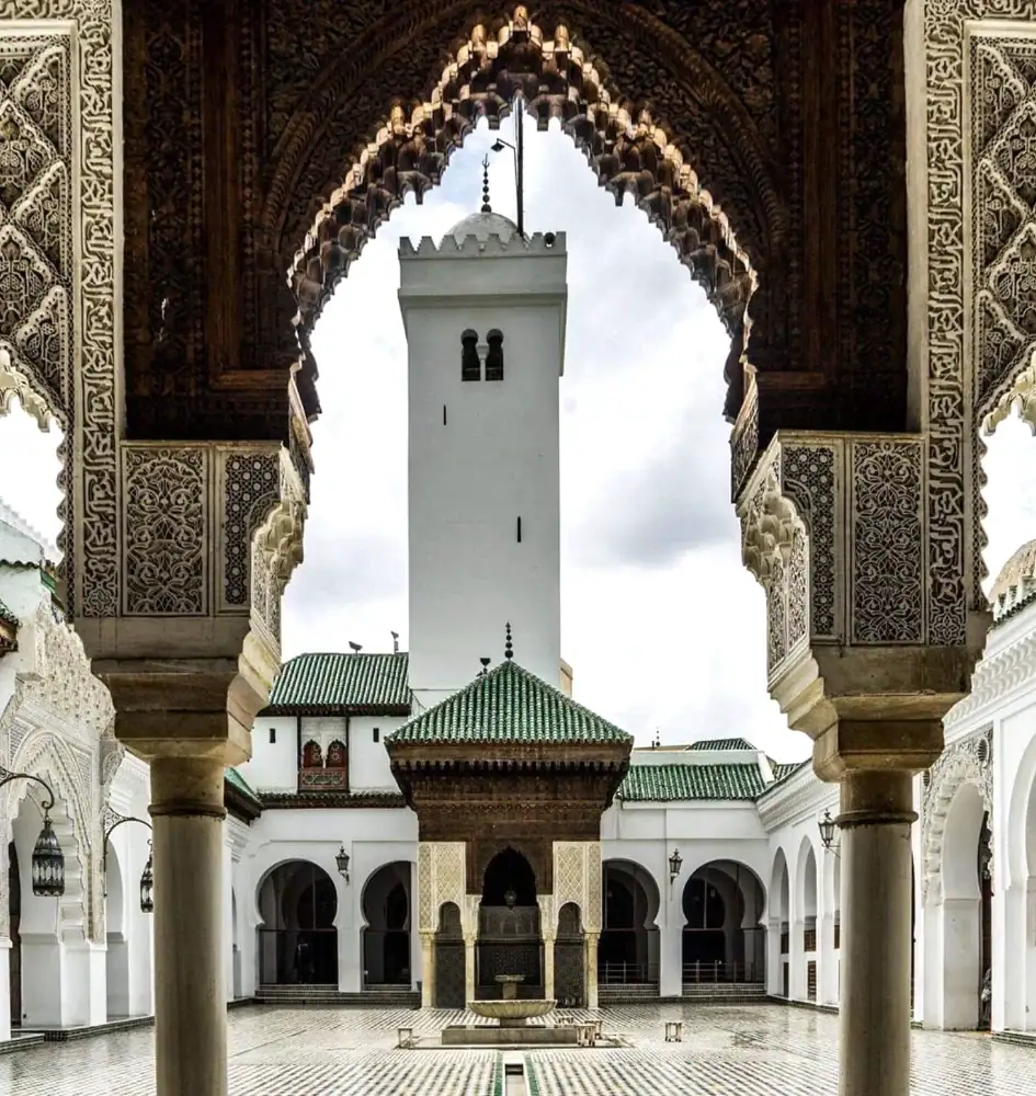 Vue depuis une arche sculptée sur la cour de l'université Al Qarawiyyin à Fès, mettant en valeur le minaret blanc, la fontaine centrale au toit de tuiles vertes et l'architecture arabo-andalouse.