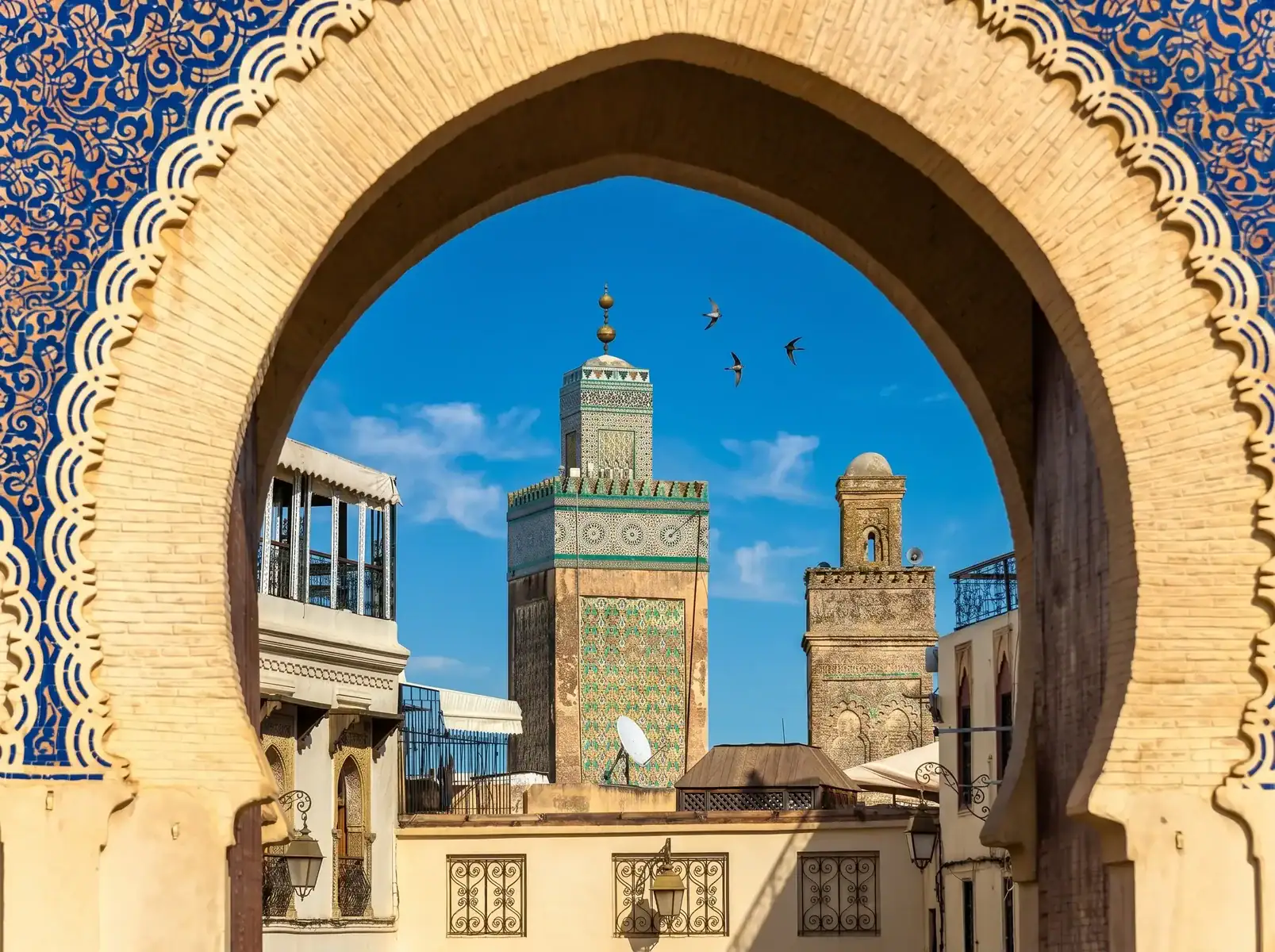 vue-bab-boujloud-medersa-bou-inania-fes Archway with intricate beige bricks frames a turquoise-tiled minaret against a blue sky, with birds flying nearby.