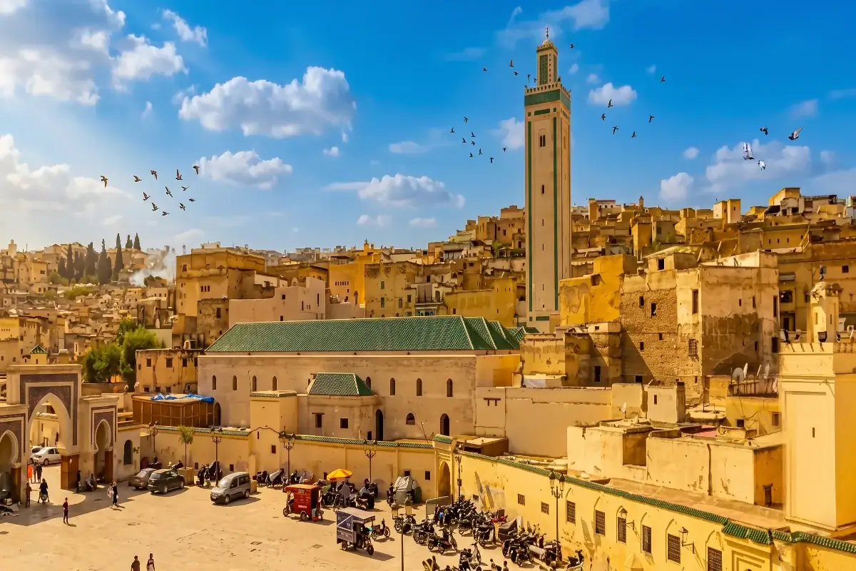 Vue panoramique sur la place animée de R'cif à Fès au Maroc, avec la mosquée au toit de tuiles vertes, son grand minaret et les habitations traditionnelles de la médina sous un ciel bleu avec des oiseaux.