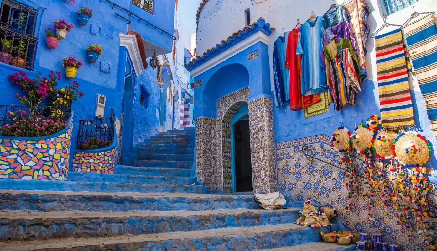 Zaouïa Ressounia à Chefchaouen Entrée de la Zaouïa Ressounia dans une ruelle bleue typique de Chefchaouen, avec des mosaïques traditionnelles, des vêtements artisanaux suspendus et des chapeaux de paille colorés.