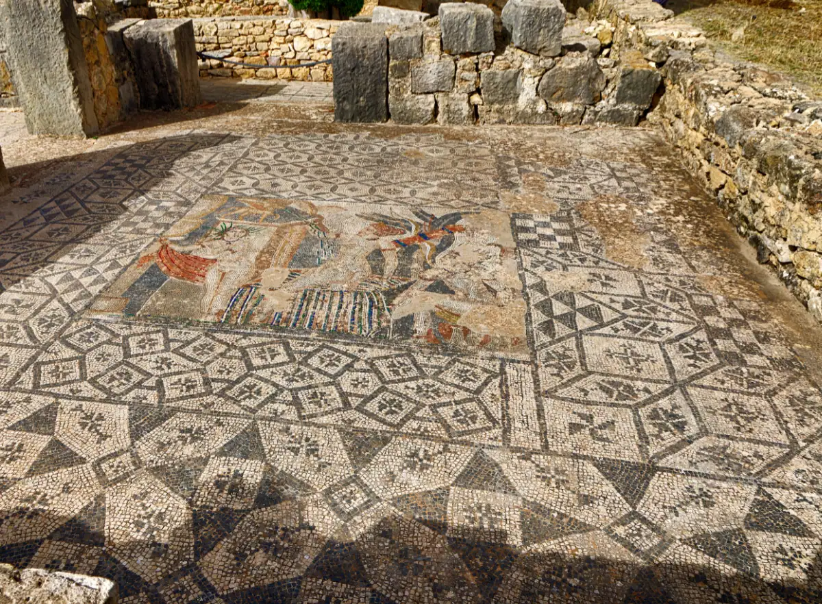 Mosaïque de sol antique présentant des motifs géométriques noirs et blancs et une scène centrale polychrome, entourée de murs en pierre en ruine sur le site archéologique de Volubilis au Maroc.