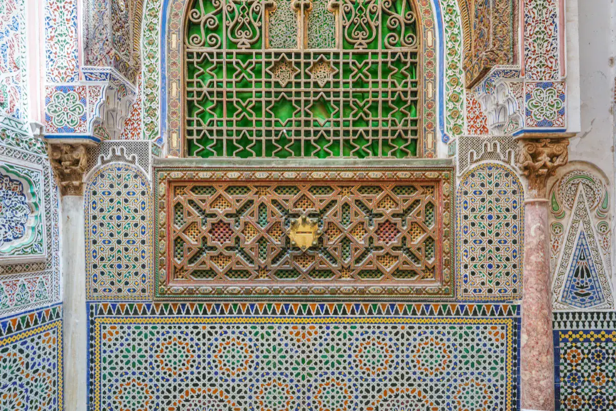 Zellige et Artisanat Marocain : Mosquée Moulay Idriss II, Fès Détail architectural complexe de la mosquée de Moulay Idriss II à Fès au Maroc. L'image montre des mosaïques traditionnelles en zellige aux motifs géométriques colorés, surmontées de panneaux en bois richement sculptés abritant une fente à aumône en laiton, et d'une grille traditionnelle verte, encadrés par des sculptures en stuc et une colonne en marbre.